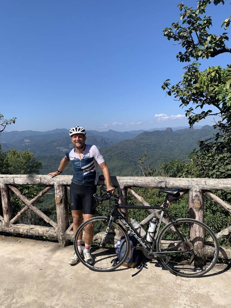 A cyclist and his bike with bright blue skies and forested mountains in the background in Mae Hong Son